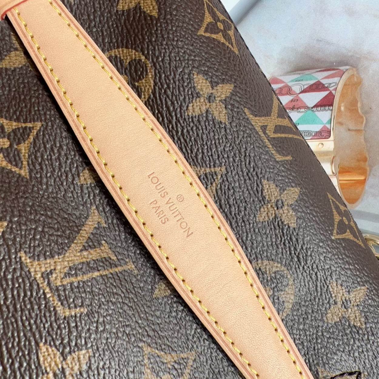 Close-up of a Louis Vuitton monogram canvas bag with a beige leather strap, showing the embossed 'Louis Vuitton Paris' text on the leather and part of a gold-tone clasp.