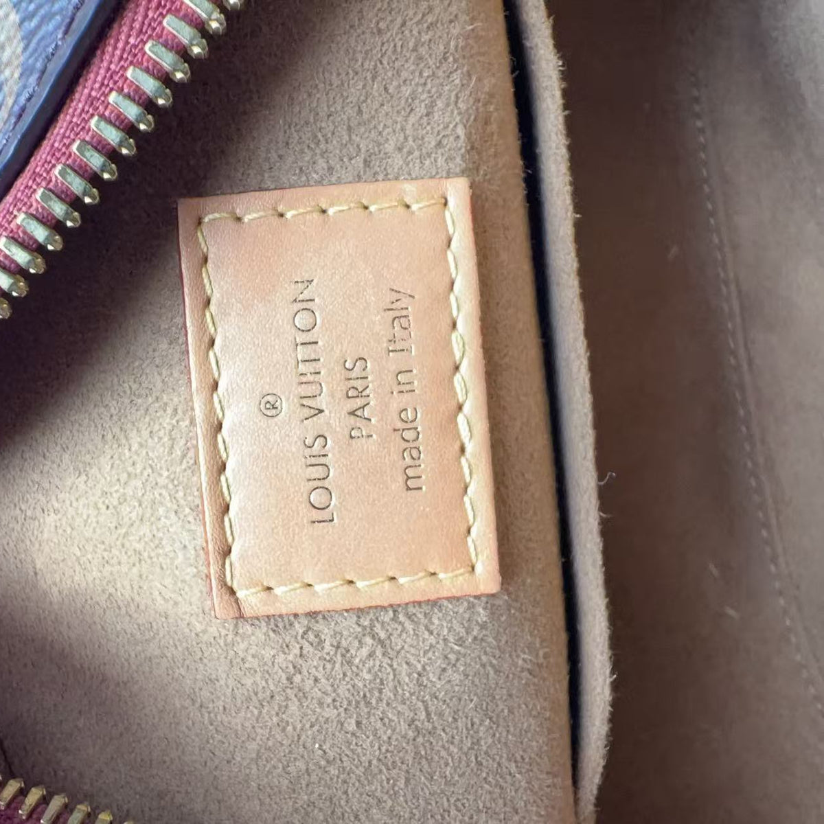 Close-up of the interior of a Louis Vuitton bag, showing a tan leather patch with 'LOUIS VUITTON PARIS made in Italy' printed on it, a gold zipper, and a blue patterned fabric edge.
