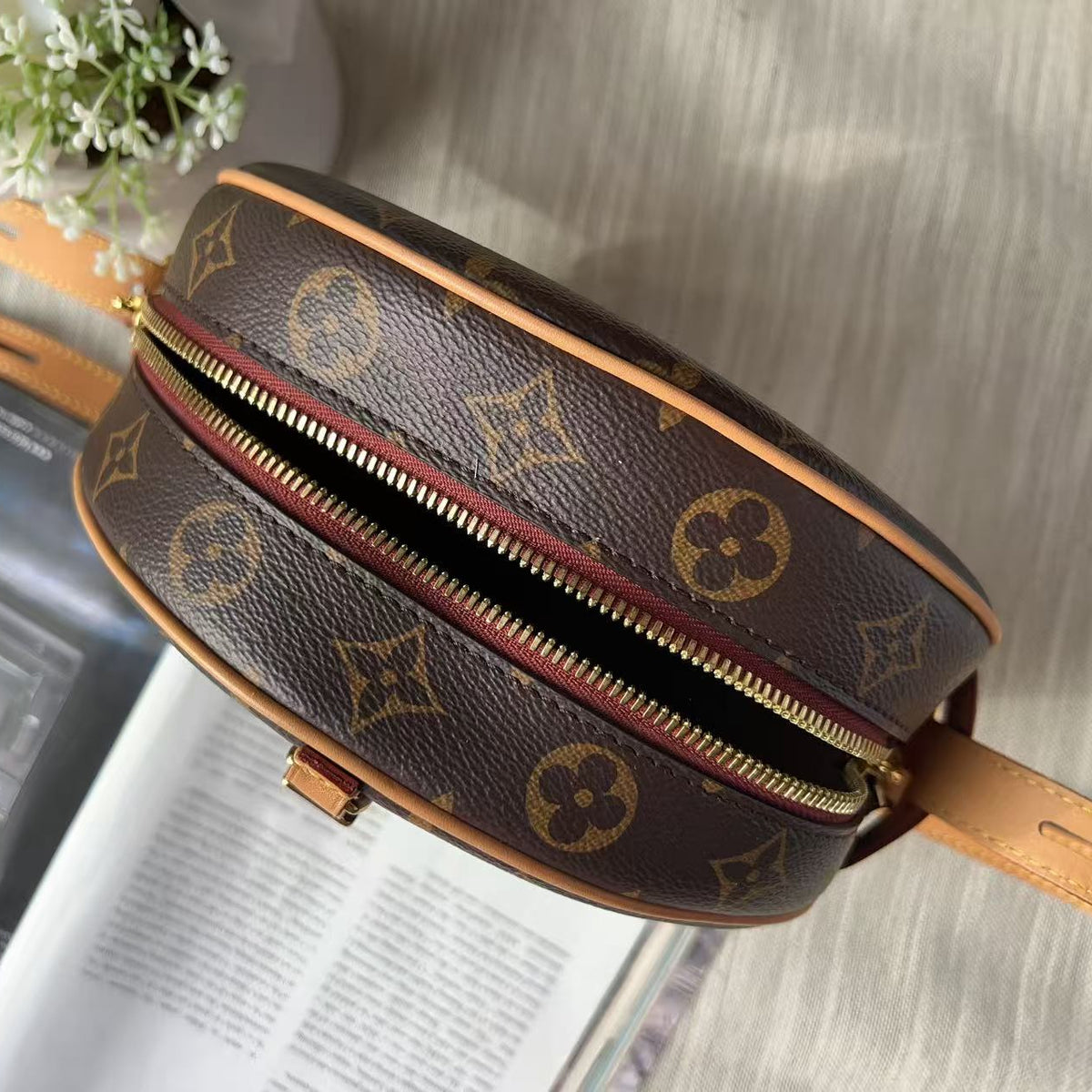 A Louis Vuitton monogram canvas round crossbody bag with a gold-tone zipper, shown in a styled flat-lay setting with a magazine and white flowers in the background.