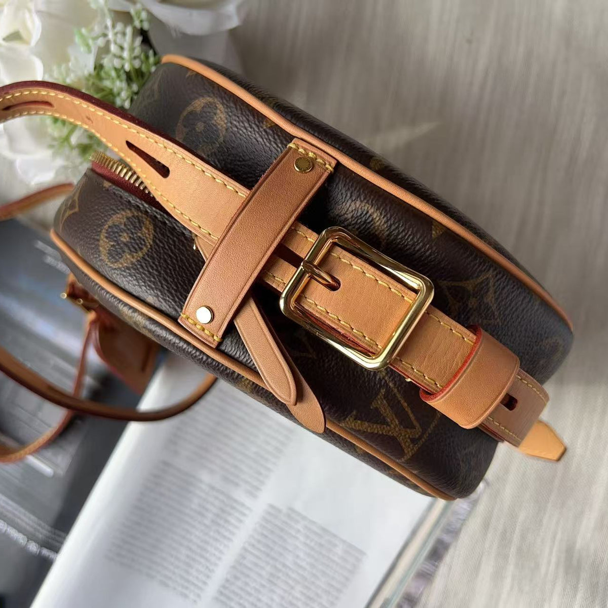 A Louis Vuitton monogram canvas round shoulder bag with tan leather trim and a gold-tone buckle, shown resting on an open magazine over a light gray surface.