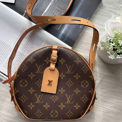 Louis Vuitton monogram canvas shoulder bag in a round, slightly asymmetrical shape, featuring tan leather trim, a matching leather hangtag with an LV logo, and a long tan shoulder strap, styled on a light textured fabric surface with books and a small plant.