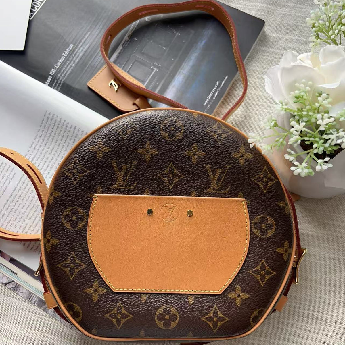 A Louis Vuitton monogrammed oval shoulder bag with a tan leather center panel, styled on a light gray textured fabric with books and white flowers.