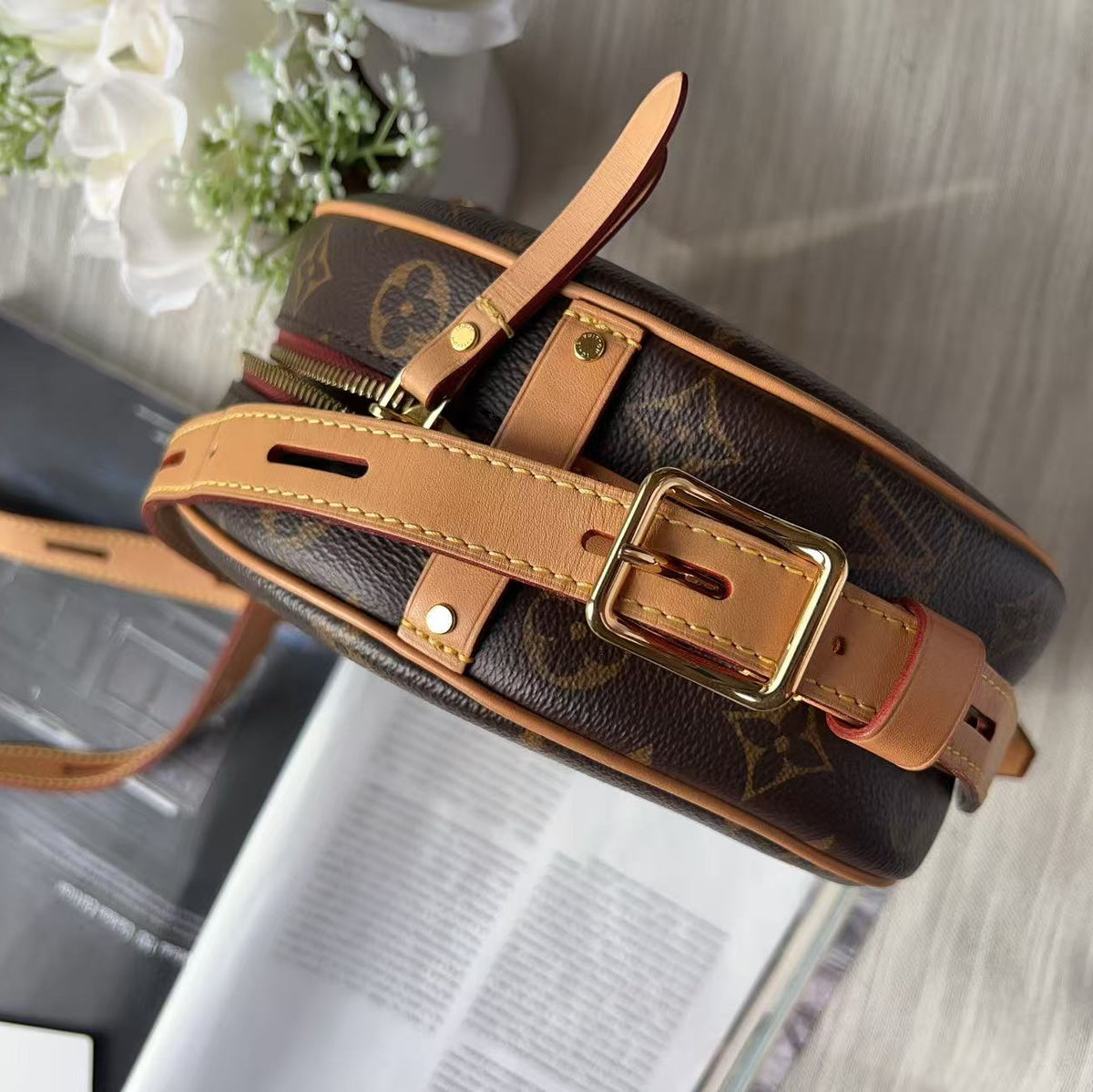 Louis Vuitton monogram canvas shoulder bag with tan leather straps and gold-tone hardware, photographed on a light wood surface with a magazine and white flowers as props.