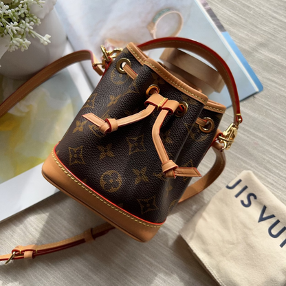 A Louis Vuitton monogram canvas bucket bag with tan leather trim and drawstring closure, styled on a light wood surface with a dust bag and magazine in the background.