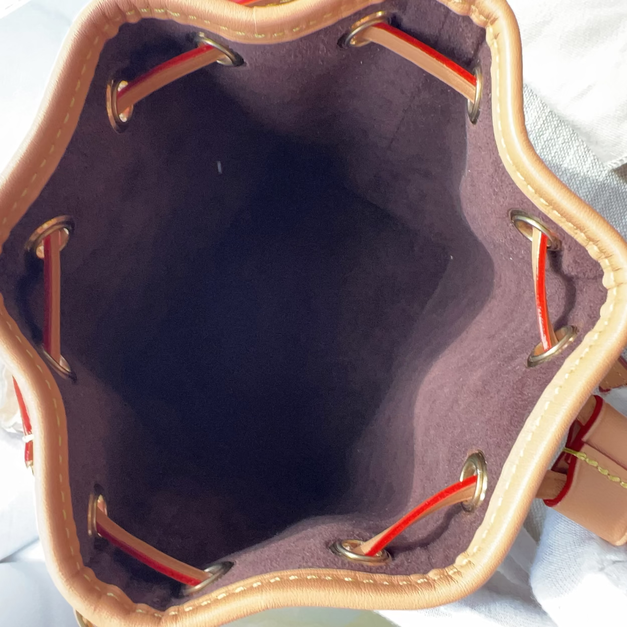 Top-down view of a bucket bag's interior, showing a dark plum purple velvet lining, tan leather trim, and red drawstring cords threaded through brass grommets.