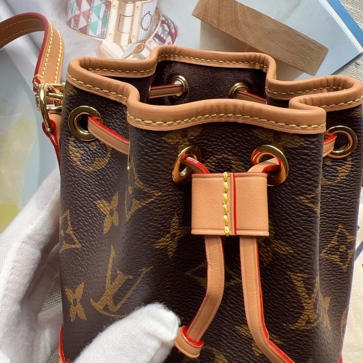 A close-up of a Louis Vuitton monogram canvas bucket bag with tan leather trim and gold-tone hardware, shown with a white-gloved hand holding it over a marble surface with jewelry props nearby.