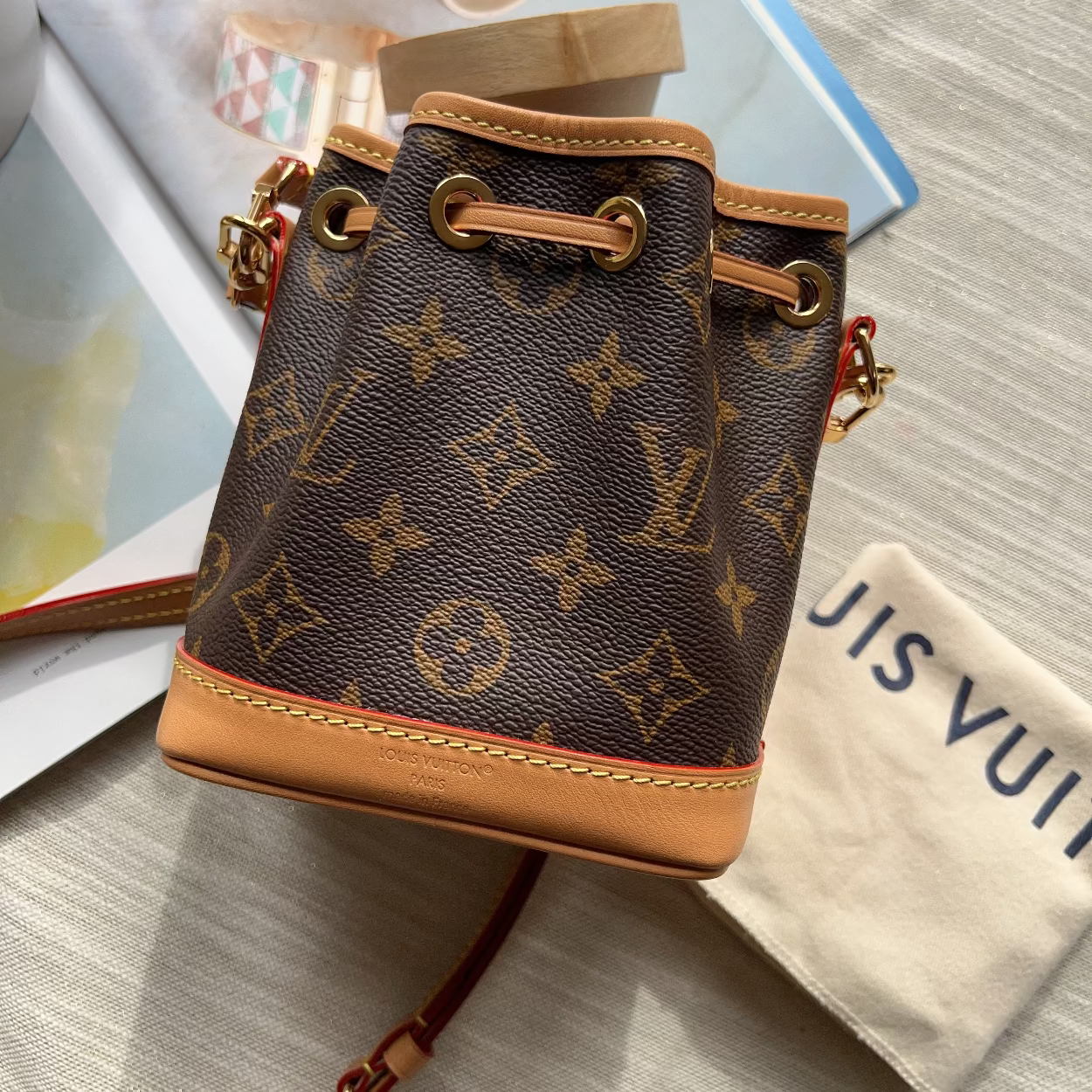 A Louis Vuitton monogram canvas bucket bag with tan leather trim and gold-tone hardware, styled on a light fabric surface with a branded dust bag and magazine pages nearby.
