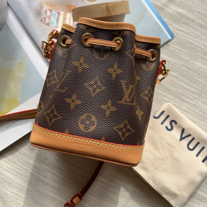 A Louis Vuitton monogram canvas bucket bag with tan leather trim and gold-tone hardware, styled on a light fabric surface with a branded dust bag and magazine pages nearby.