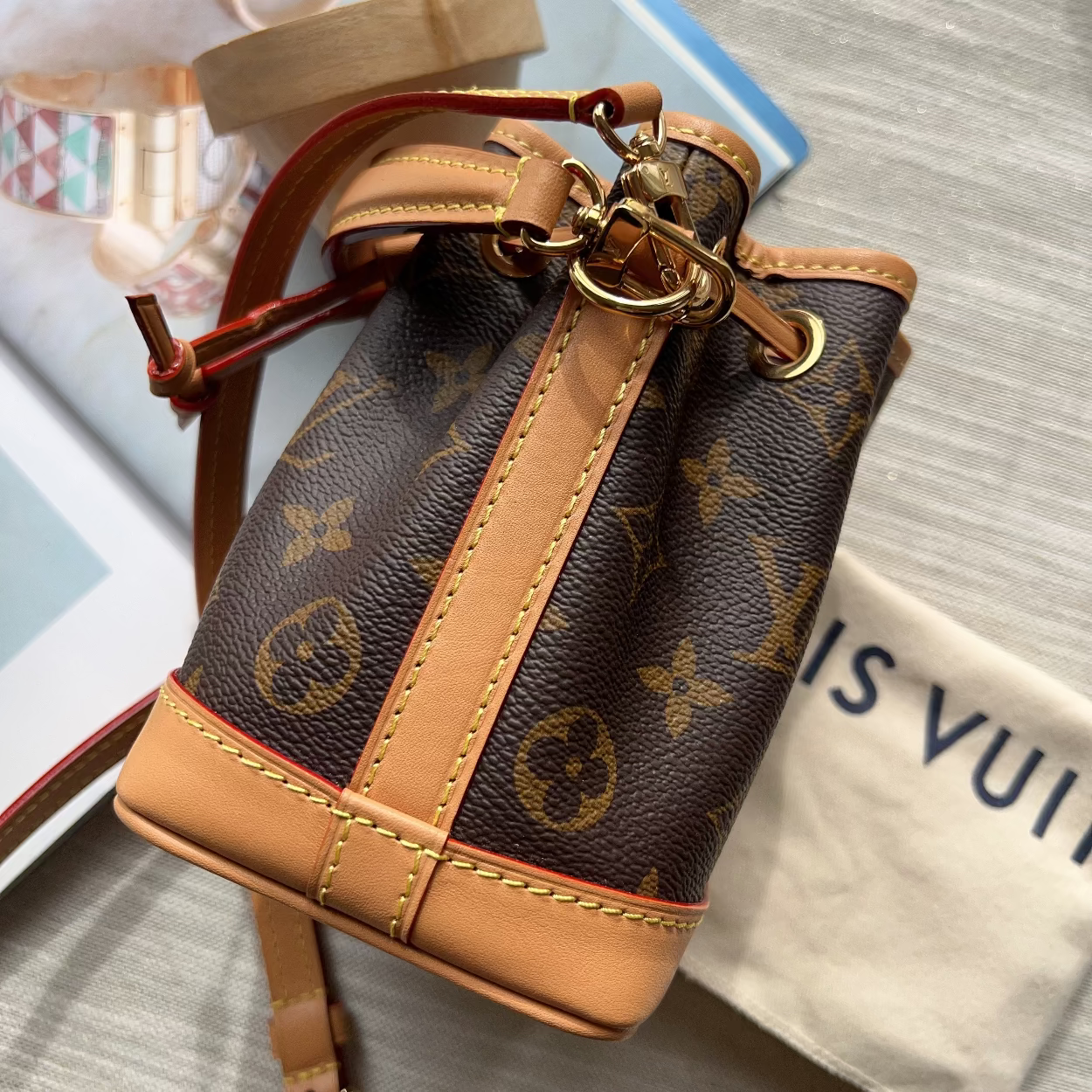 A Louis Vuitton monogram canvas bucket bag with tan leather trim and gold-tone hardware, shown on a light gray textured surface with a dust bag and accessories in the background.