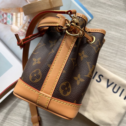 A Louis Vuitton monogram canvas bucket bag with tan leather trim and gold-tone hardware, shown on a light gray textured surface with a dust bag and accessories in the background.