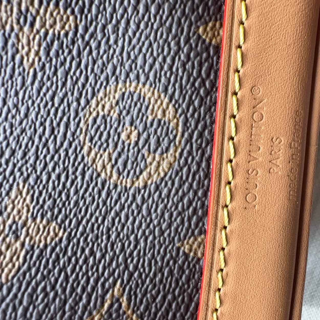 Close-up of a Louis Vuitton monogram canvas item with tan leather trim and yellow stitching, showing embossed 'LOUIS VUITTON S.A.