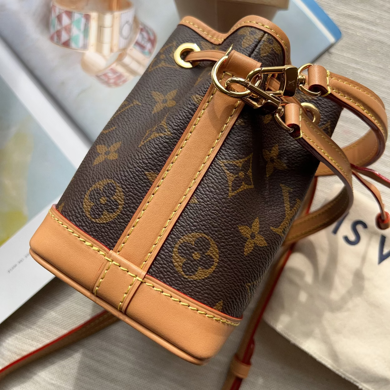 A Louis Vuitton monogram canvas bucket bag with tan leather trim and gold-tone hardware, shown resting on a light-colored surface with a branded dust bag and accessories in the background.