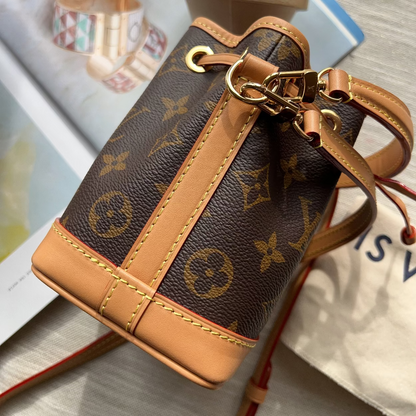A Louis Vuitton monogram canvas bucket bag with tan leather trim and gold-tone hardware, shown resting on a light-colored surface with a branded dust bag and accessories in the background.