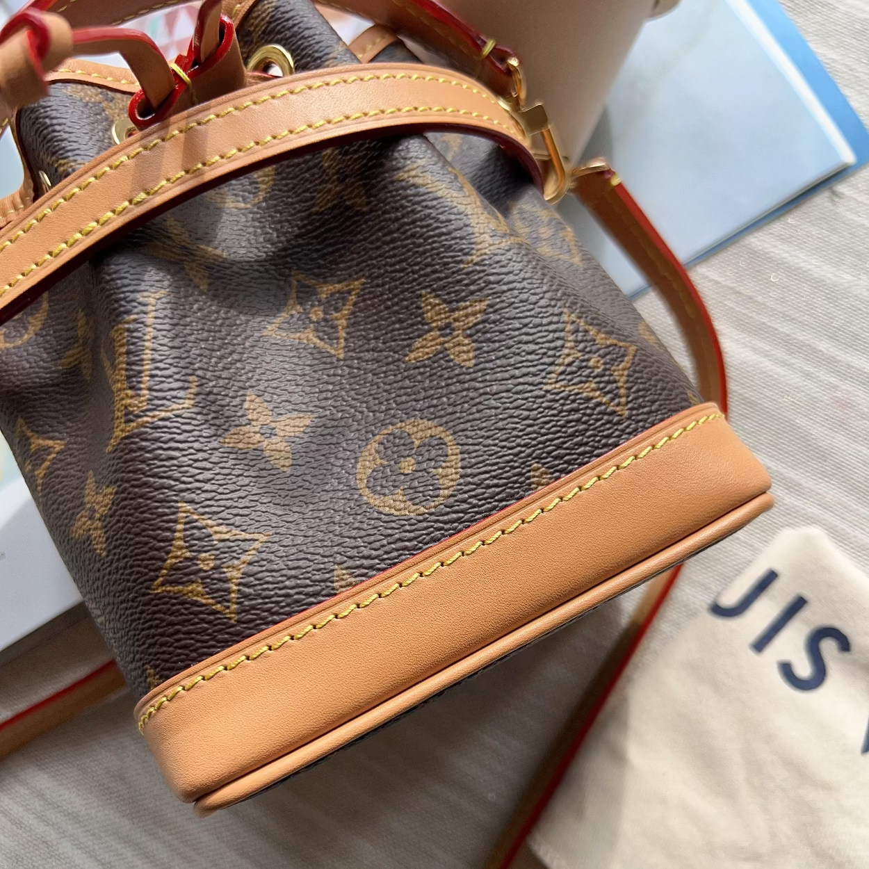 Louis Vuitton monogram canvas bucket bag with tan leather trim and a red drawstring, shown on a light wood surface next to a dust bag.