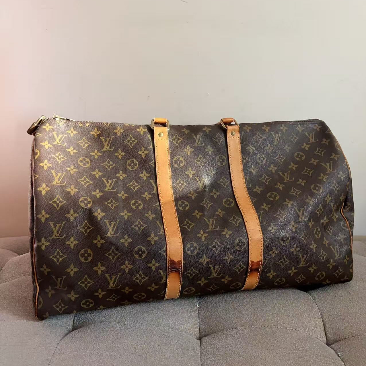A Louis Vuitton monogram canvas duffel bag with tan leather trim, resting on a gray tufted sofa against a plain light wall.