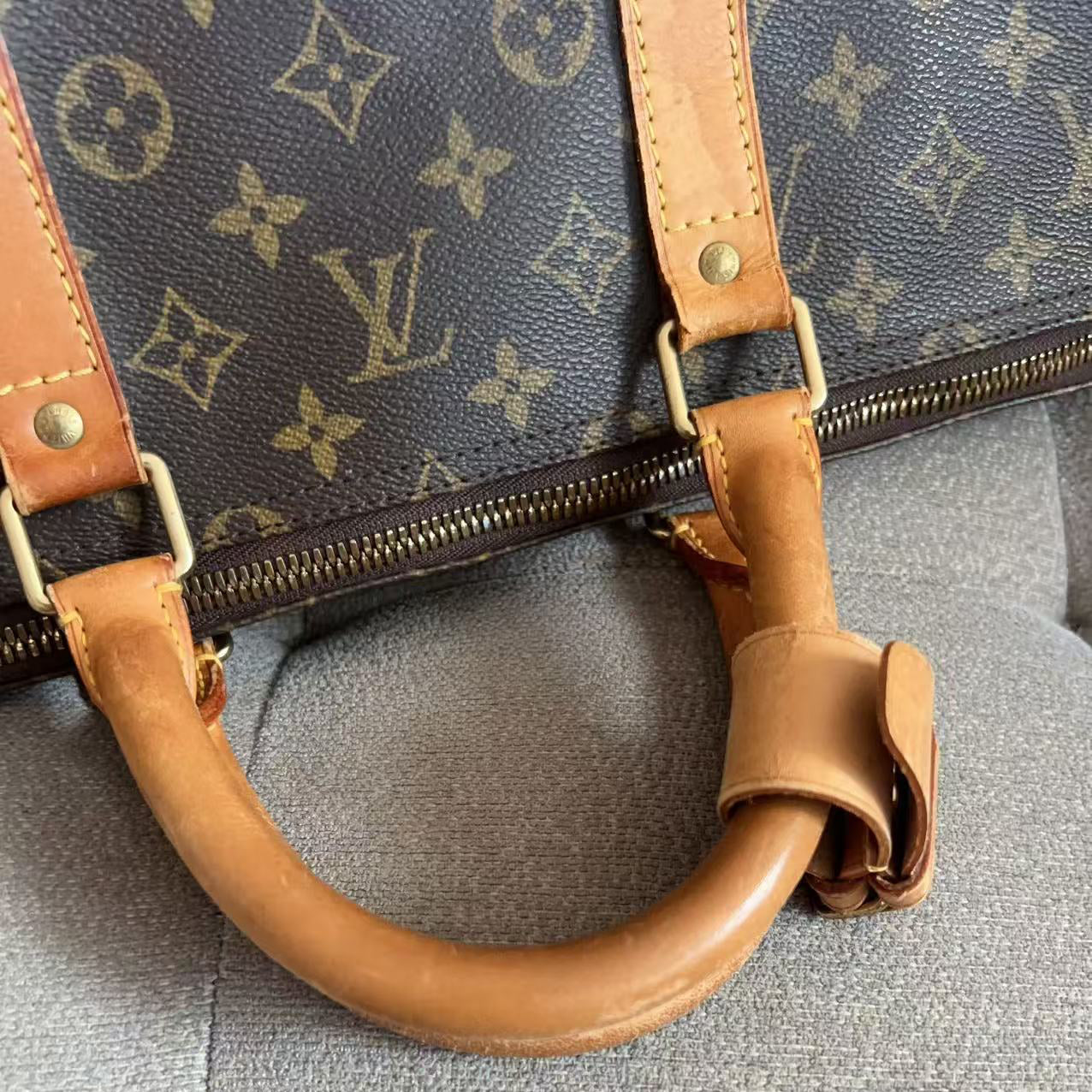 Close-up of a Louis Vuitton monogram canvas handbag with tan leather handles and hardware, resting on a gray textured surface.