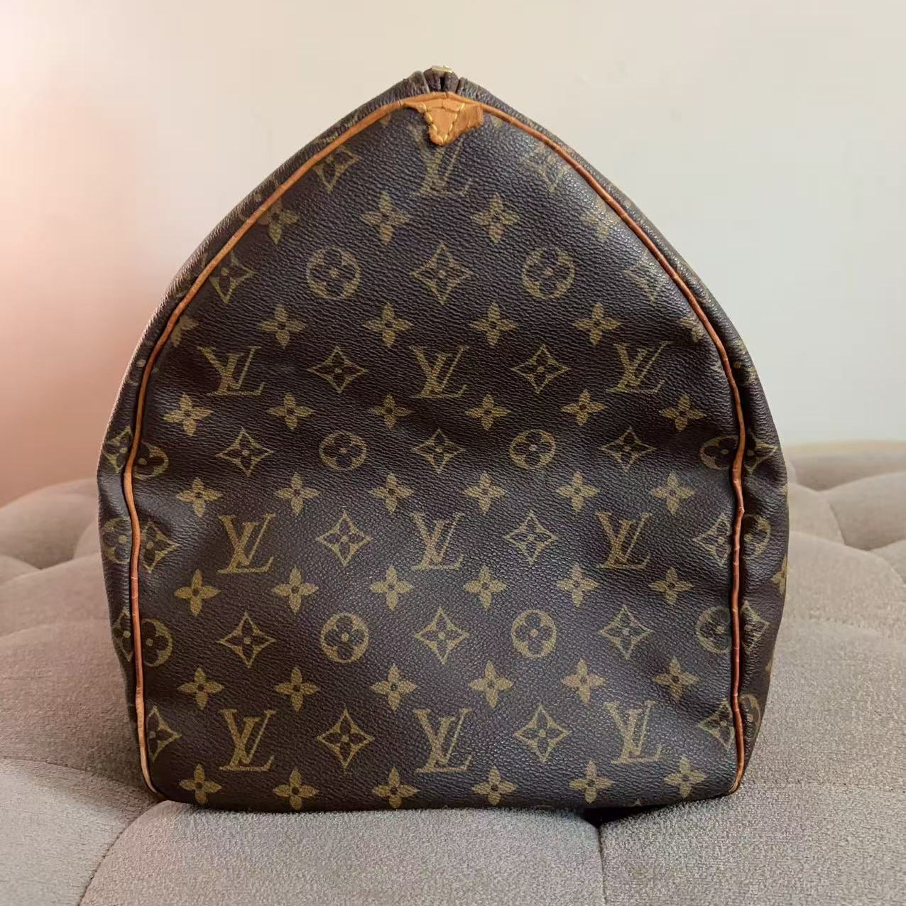 A Louis Vuitton monogram canvas duffel bag in dark brown with tan leather trim, resting on a light gray tufted chair against a plain off-white wall.