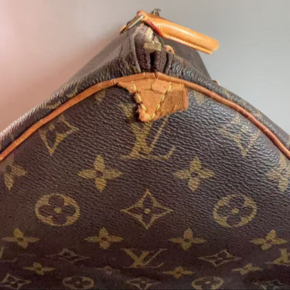 Close-up of a Louis Vuitton monogram canvas bag corner showing the brown coated canvas with tan monogram print, a tan leather trim, and a tan zipper pull against a plain light background.