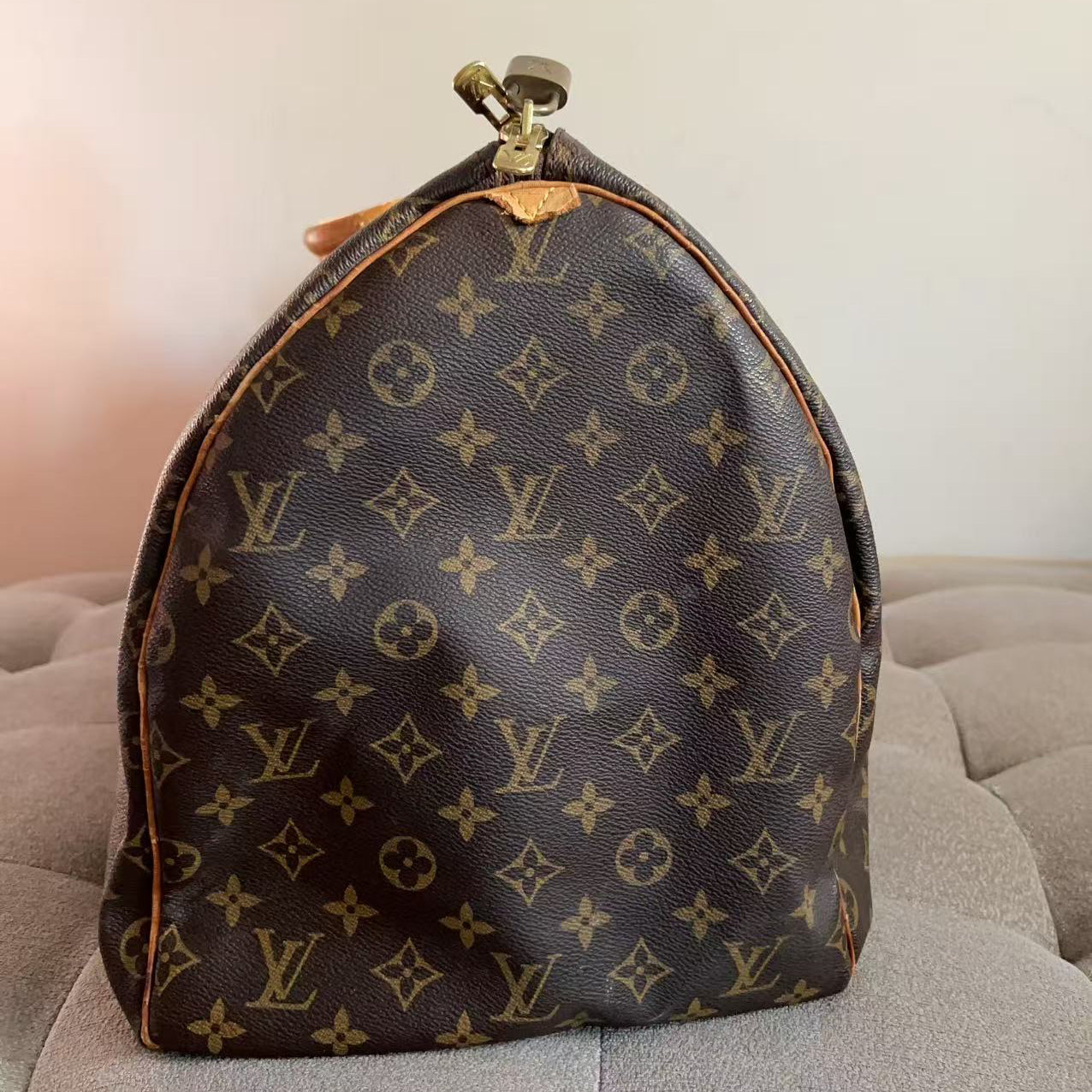 A Louis Vuitton monogram canvas dust bag or small bag with a rounded top and gold-tone zipper, resting on a gray tufted surface against a plain light wall.