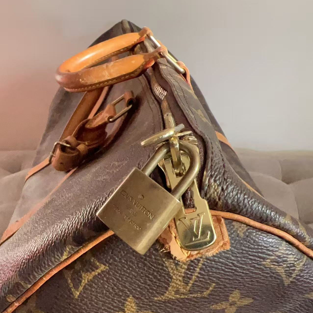 Close-up of a Louis Vuitton monogram duffel bag in dark brown coated canvas with tan leather trim and a brass padlock hanging from the zipper.