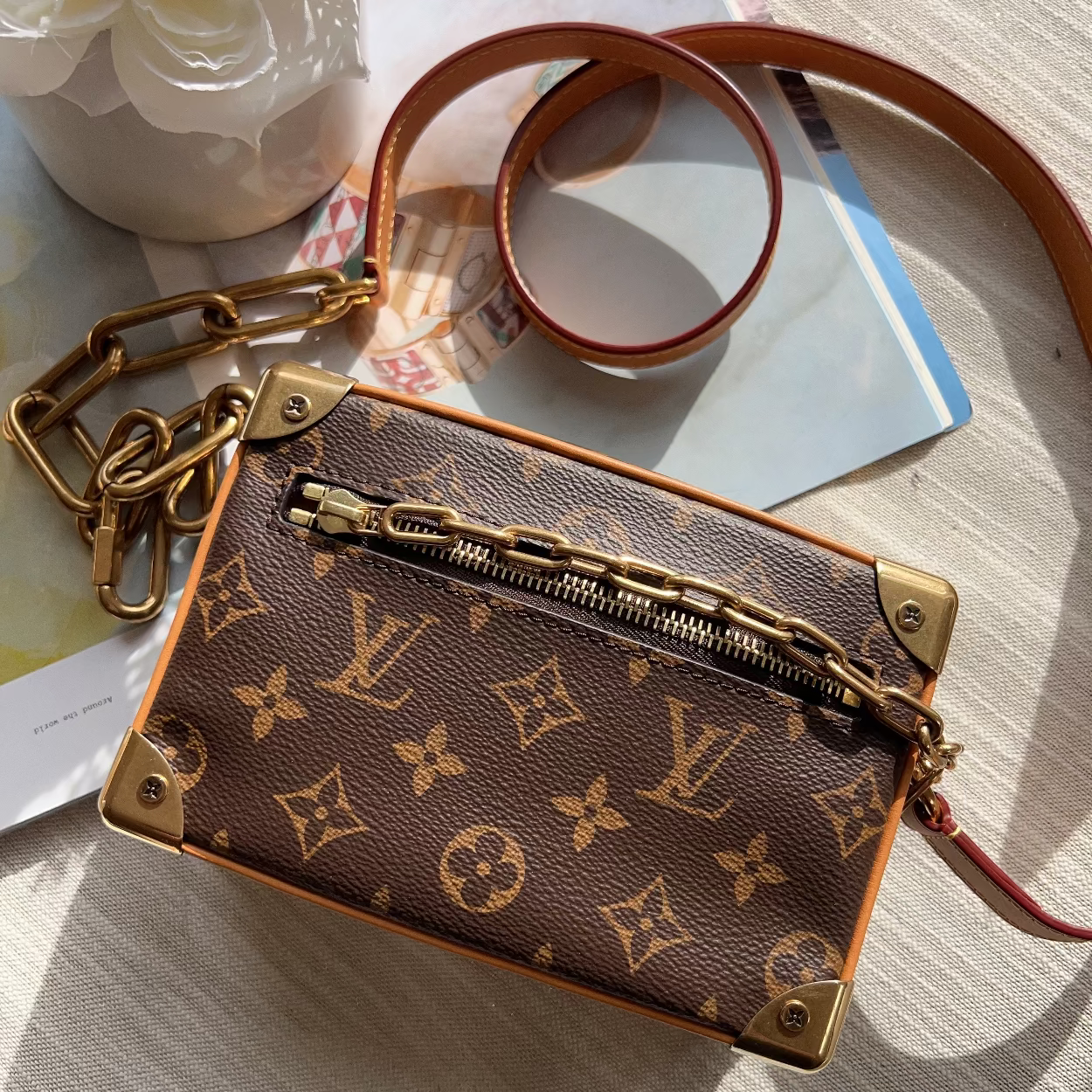 A Louis Vuitton monogram canvas mini trunk shoulder bag with gold-tone hardware, displayed on a light beige fabric background with a magazine and white flowers as props.