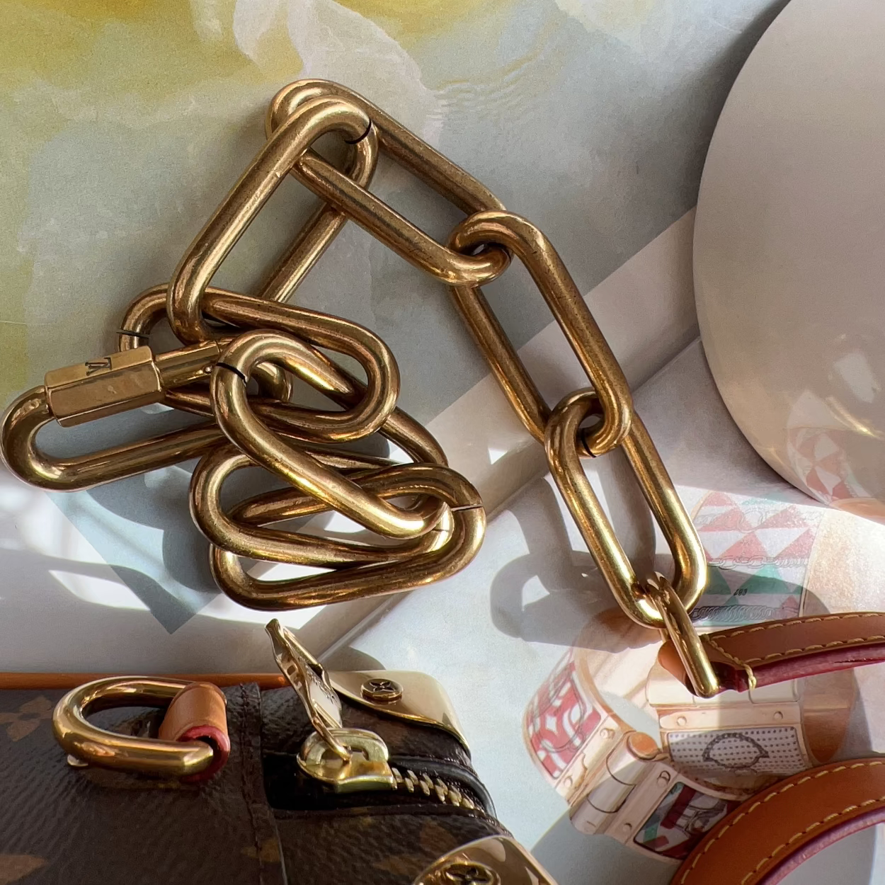 A close-up of a chunky gold-tone metal chain strap draped over a brown Louis Vuitton monogram bag, with part of a white and yellow patterned surface and a white plate visible in the background.