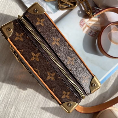 A close-up of a Louis Vuitton monogram canvas zippered case or organizer, showing the brown coated canvas with tan floral motifs, gold-tone hardware, and a light brown leather trim, photographed on a light fabric surface with a magnifying glass and strap in the background.