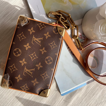 A Louis Vuitton monogram canvas phone pouch with gold-tone hardware and a tan leather strap, styled on a light fabric surface with books and a white flower vase in the background.