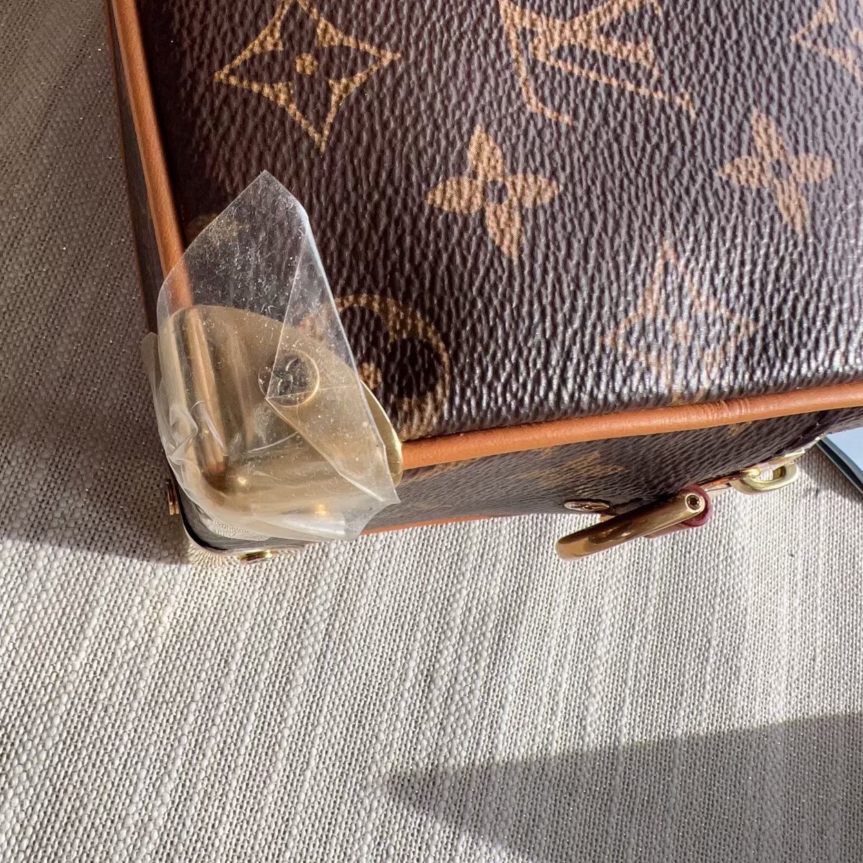 A close-up of a brown Louis Vuitton monogram canvas bag corner with gold-tone hardware and a clear protective plastic film partially peeled back, resting on a textured beige fabric surface.