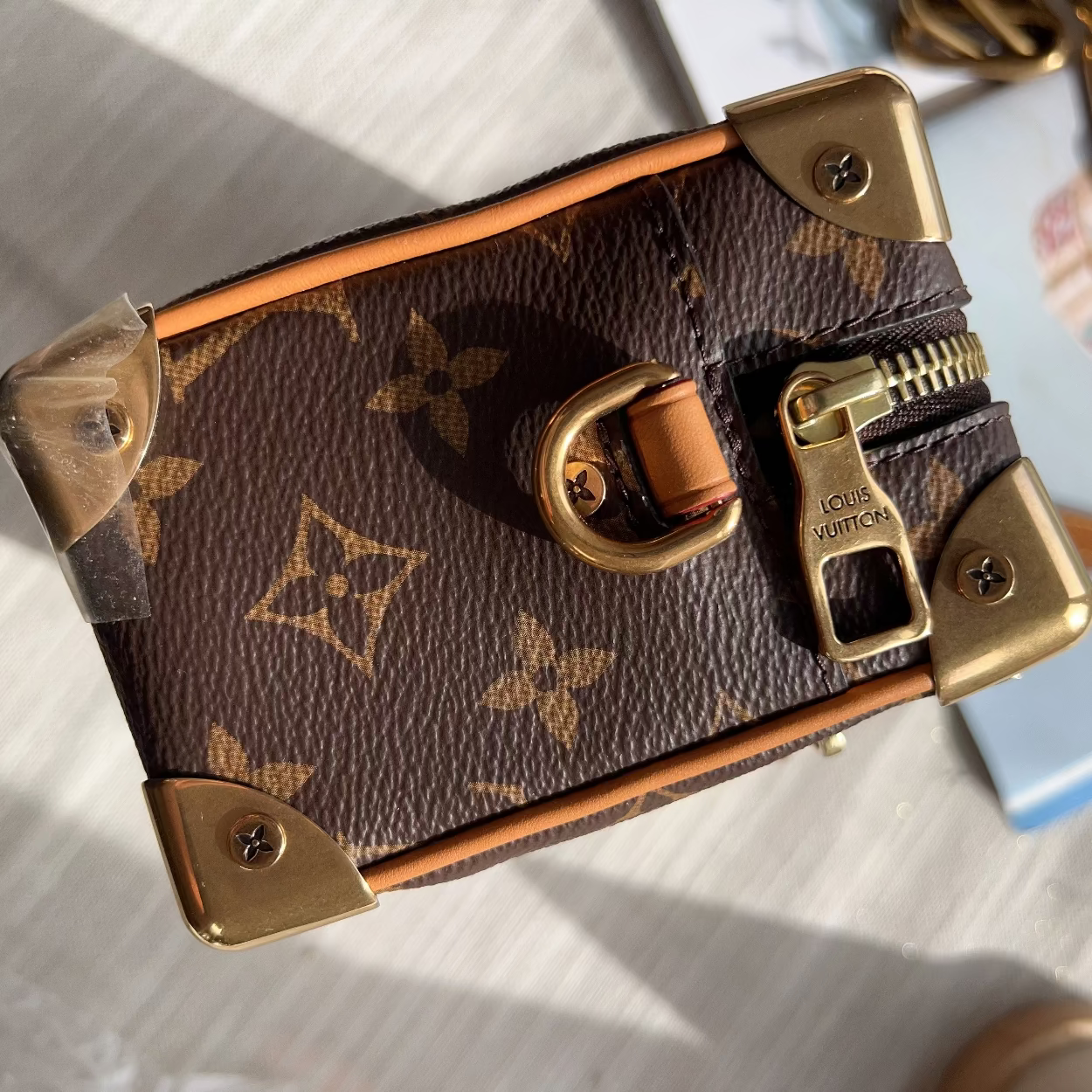 A Louis Vuitton miniature trunk bag in brown monogram canvas with gold-tone hardware, shown in a close-up angled view on a light surface.