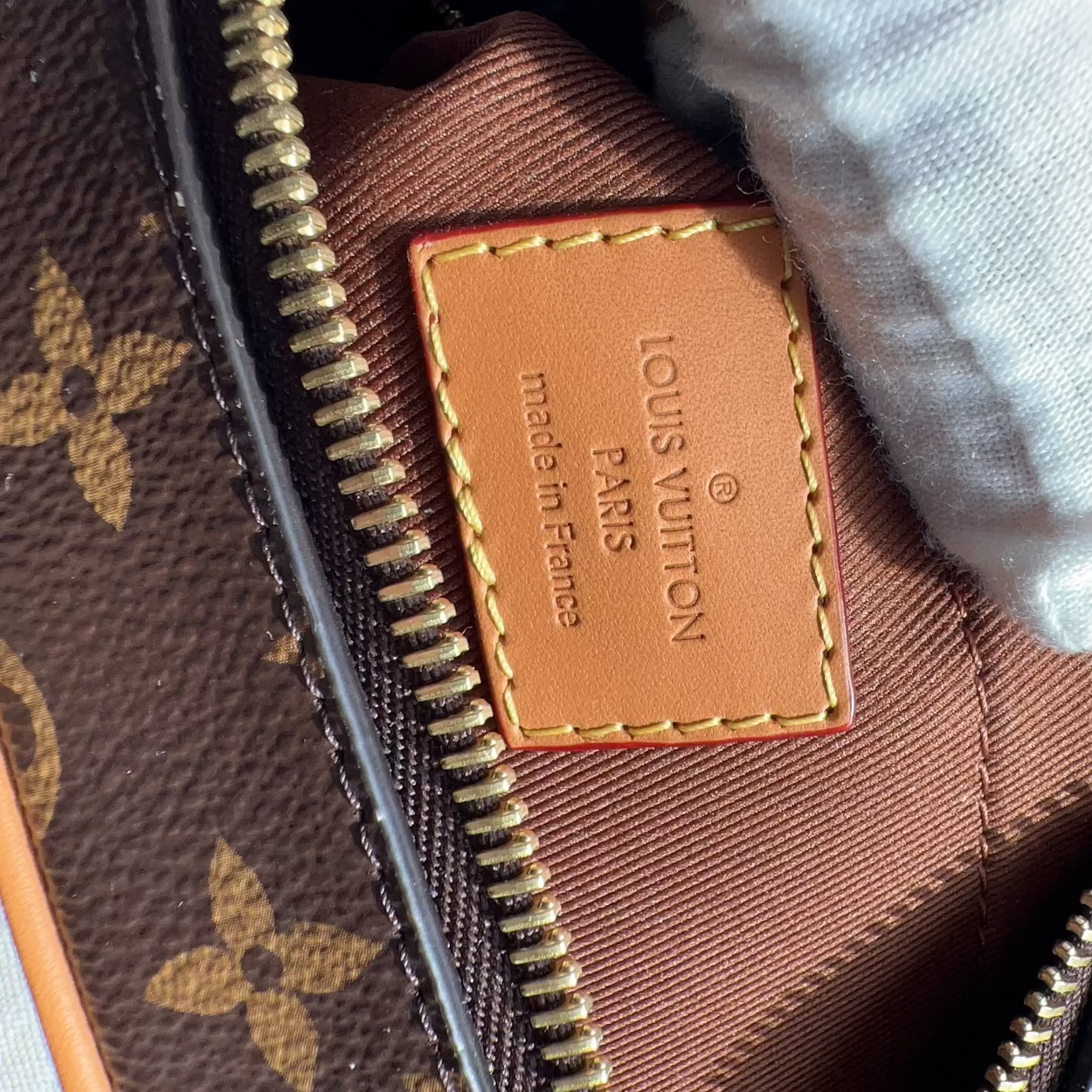 Close-up of a Louis Vuitton monogram bag interior showing a tan leather patch with 'LOUIS VUITTON PARIS made in France' text, gold zipper, brown fabric lining, and a white textured fabric or glove at the top right.
