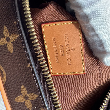 Close-up of a Louis Vuitton monogram bag interior showing a tan leather patch with 'LOUIS VUITTON PARIS made in France' text, gold zipper, brown fabric lining, and a white textured fabric or glove at the top right.