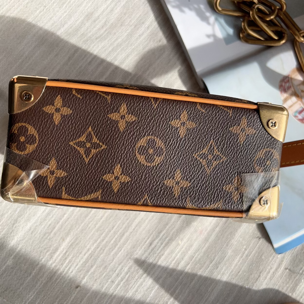 A close-up of a Louis Vuitton monogram canvas handbag corner with gold-tone metal hardware, shown on a light textured surface with part of a chain strap visible.