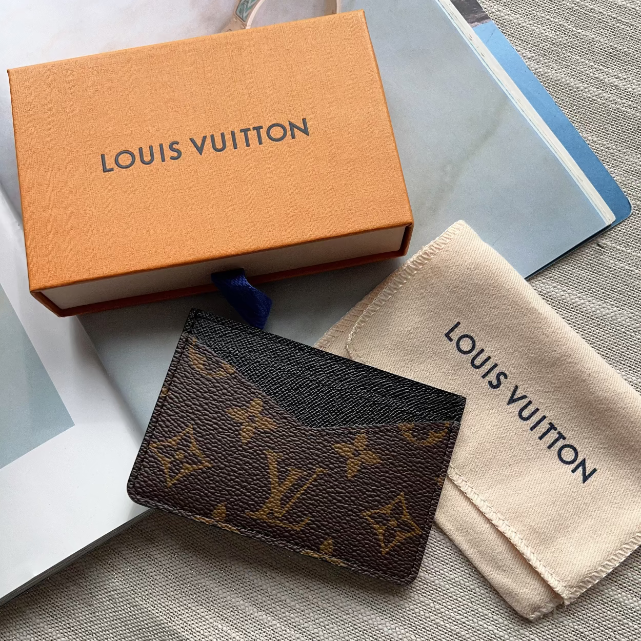 A Louis Vuitton monogram card holder or small wallet in dark brown coated canvas with gold-tone LV monogram motifs, shown with its orange Louis Vuitton box and beige dust bag on a gray textured fabric surface.
