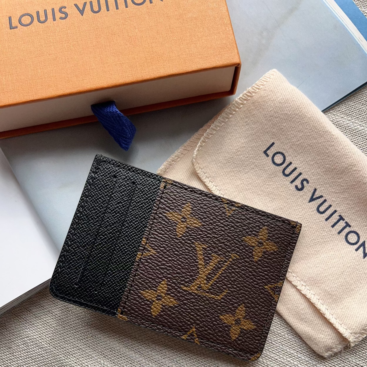 A Louis Vuitton monogram card holder wallet with a black textured leather half, displayed next to its orange box, dust bag, and notebook on a gray textured surface.