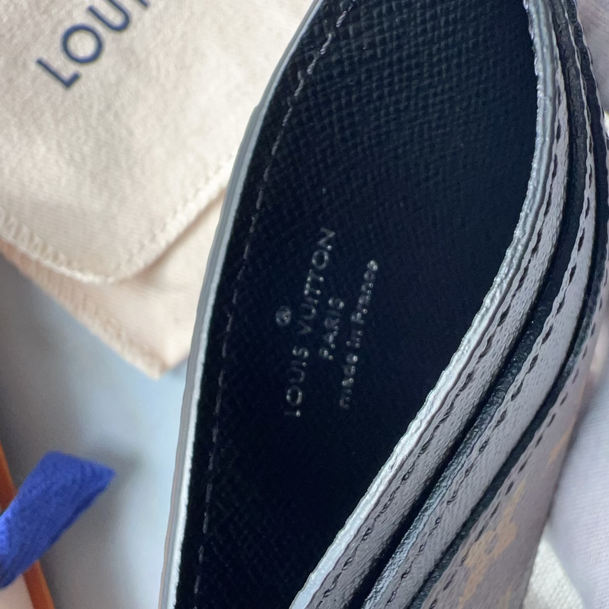 Close-up of the interior of a Louis Vuitton wallet or card case, showing the black textured Vachetta leather lining with white printed brand text and white stitching. A cream dust bag with blue Louis Vuitton lettering and part of an orange box with a blue ribbon are visible in the background.