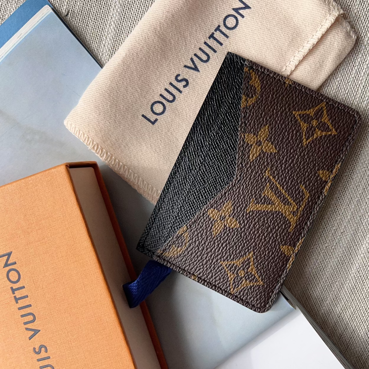 A Louis Vuitton monogram card holder or small wallet in brown coated canvas with gold-tone LV monogram print, shown with a cream dust bag and orange box on a gray fabric surface.