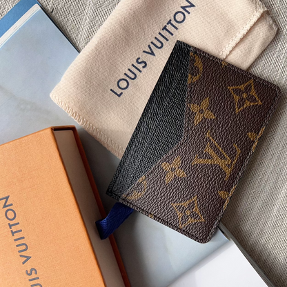 A Louis Vuitton monogram card holder or small wallet in brown coated canvas with gold-tone LV monogram print, shown with a cream dust bag and orange box on a gray fabric surface.