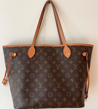 A Louis Vuitton monogram canvas tote bag with tan leather shoulder straps and trim, hanging against a plain off-white wall.