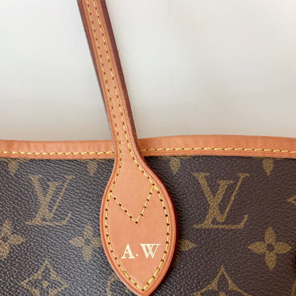Close-up of a Louis Vuitton monogram tote bag showing the brown coated canvas with tan monogram print, a tan leather handle, and a central leather tab stamped with 'A.W'.