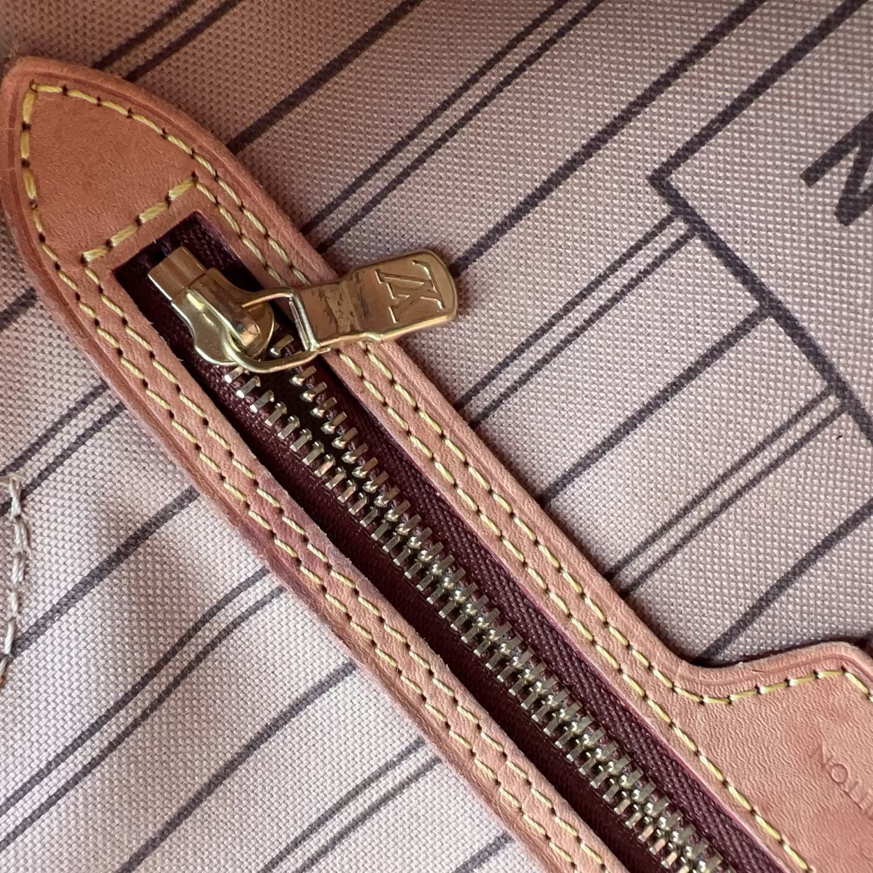 Close-up of a zipper pull tab made of tan leather with gold-tone metal hardware, shown on a striped fabric surface.