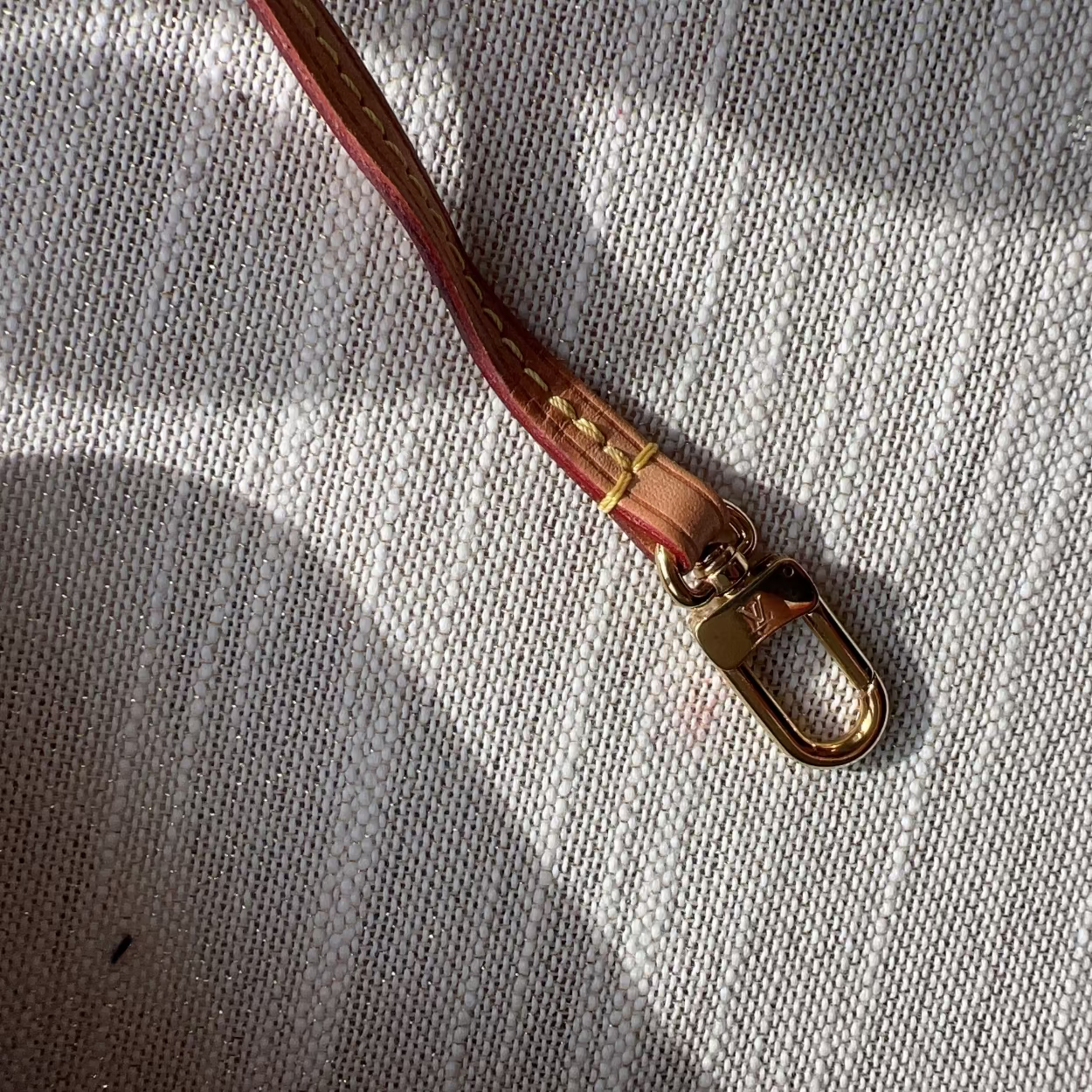 A close-up of a brown leather strap with gold-tone metal hardware, laid diagonally on a textured off-white knit fabric under dappled sunlight.