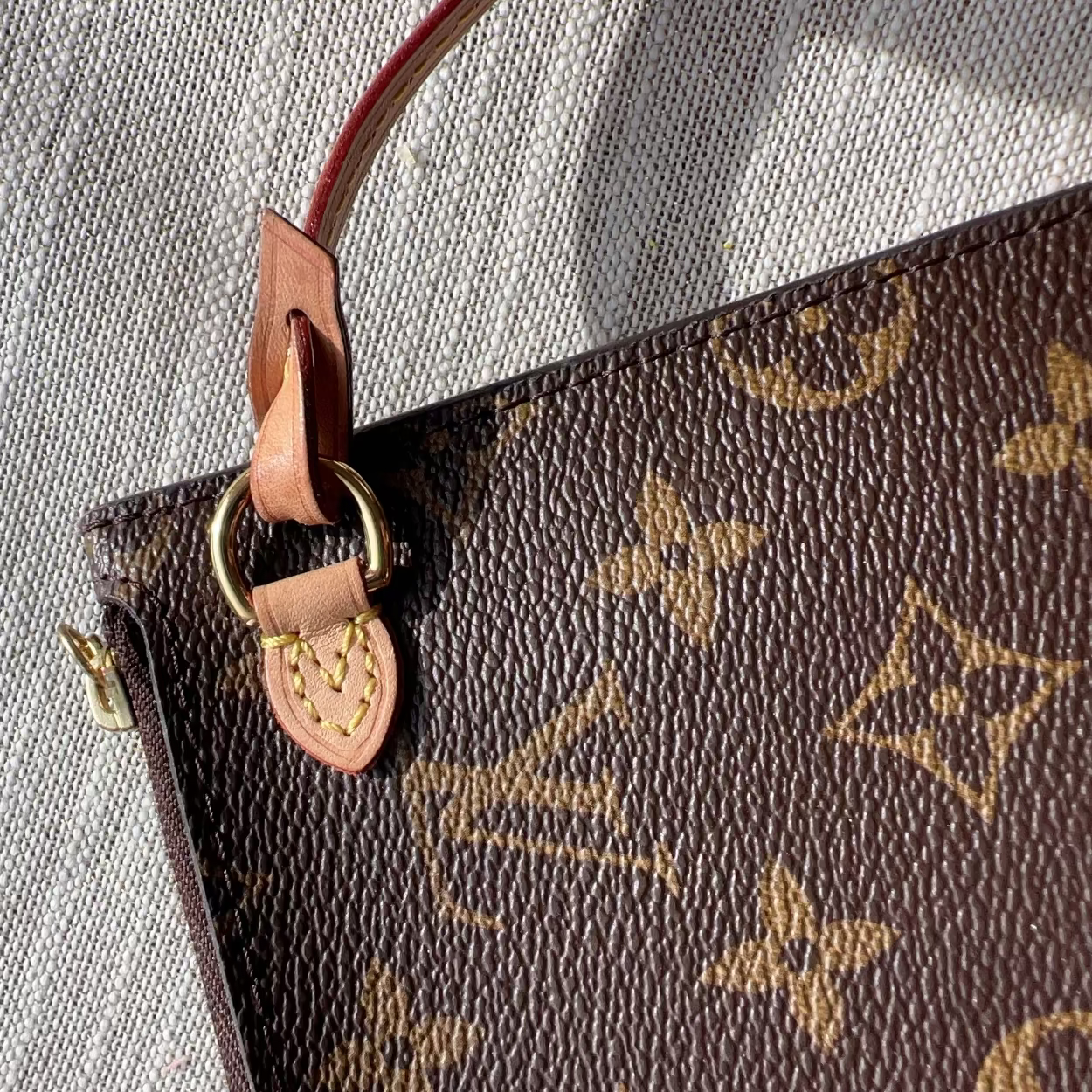 Close-up of a brown monogram handbag corner with tan leather trim, gold-tone hardware, and a textured light gray fabric background.