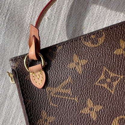 Close-up of a brown monogram handbag corner with tan leather trim, gold-tone hardware, and a textured light gray fabric background.