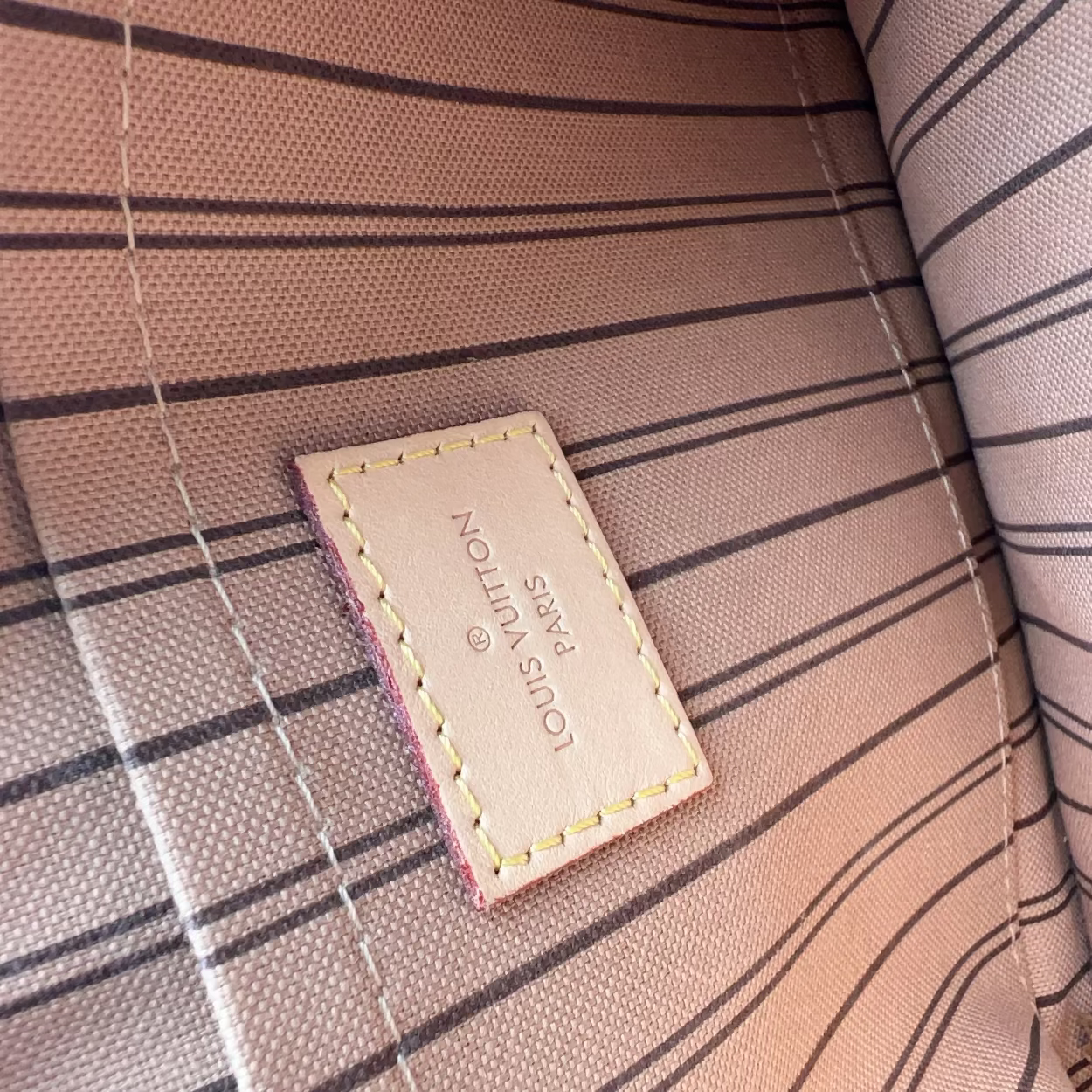 Close-up of a Louis Vuitton interior lining patch on a striped fabric surface. The patch reads 'LOUIS VUITTON PARIS' and is stitched onto a beige and brown plaid textile background.