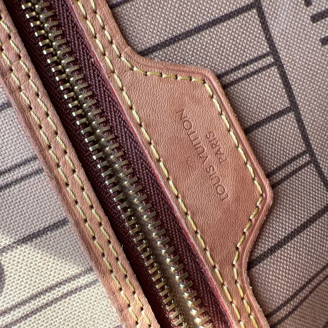 Close-up detail of a Louis Vuitton zippered pouch or wallet, showing a dusty pink leather tab with gold-tone metal hardware, contrast stitching, and the debossed brand name on a striped canvas background.