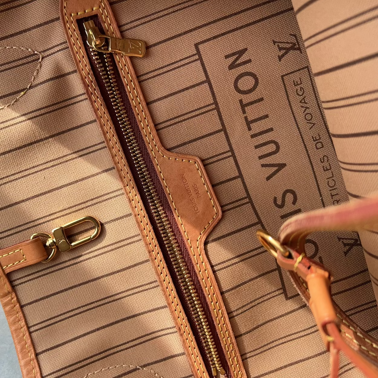 Close-up of a Louis Vuitton tote bag interior showing the striped beige canvas, tan leather trim, gold-tone zipper, and printed Louis Vuitton travel text.