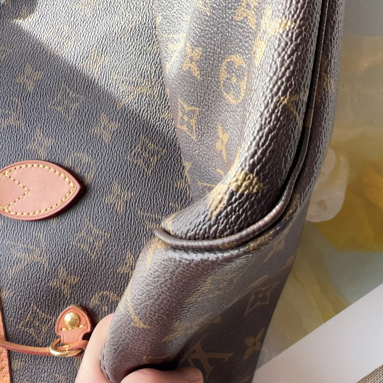 A close-up of a Louis Vuitton monogram canvas bag being held by a hand, showing the brown coated canvas with gold monogram print and a tan leather accent piece.