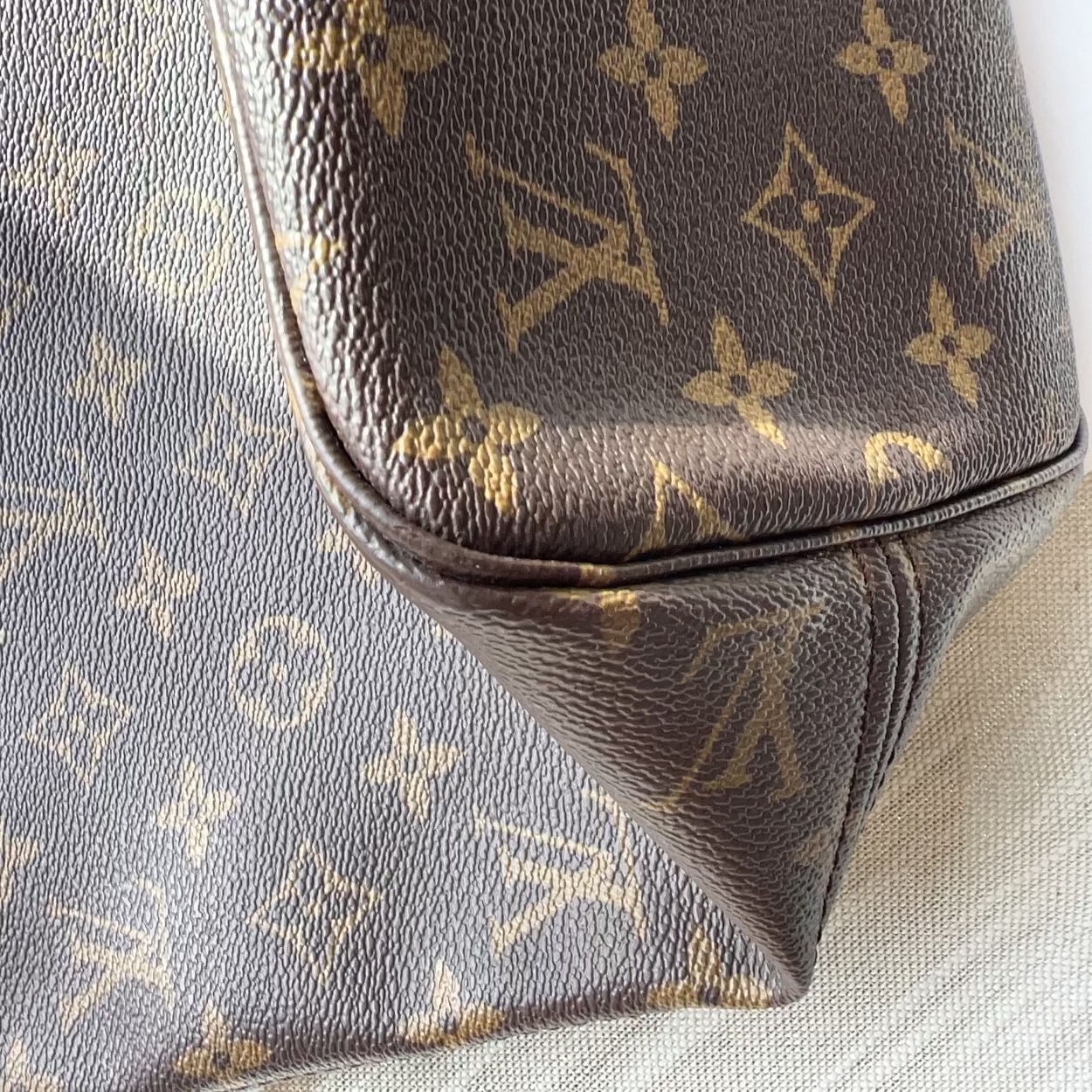 Close-up of a Louis Vuitton monogram canvas bag in dark brown and beige, showing the classic LV logo pattern, a gold-tone metal ring, and a piece of light brown leather trim on a light textured fabric background.