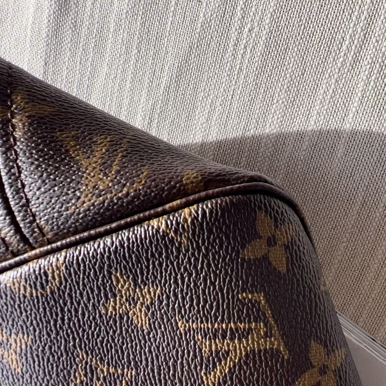 Close-up of a brown monogram handbag corner with gold-toned Louis Vuitton-style floral and LV motifs, set against a light beige textured fabric background.